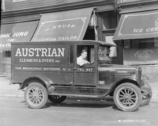 View across street towards a man sitting in the passenger seat of a truck parked in front of the J. Krupp Custom Tailor storefront. The sign on the truck reads: "Austrian Cleaners & Dyers Inc."