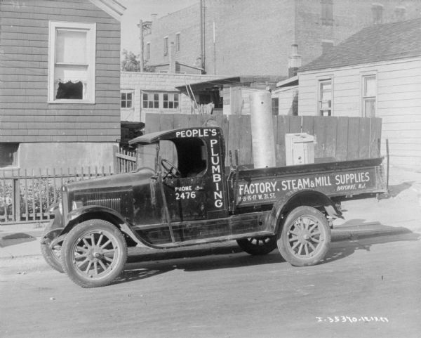 View across street towards a delivery truck parked along a curb. There is a house and garage behind a fence in the background. The sign painted on the truck reads: "People's Plumbing, Factory, Steam & Mill Supplies, Bayonne, N.J."