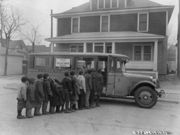 View across street towards children waiting in a line to get on a Harvester Coach bus. A sign in the window of the bus reads: "Body Built by Lang Body Company."