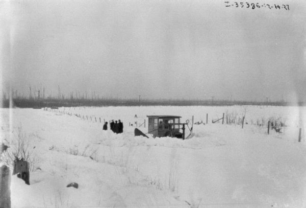 A group of men are standing near a truck with a snowplow in a rural area. There is deep snow on the ground.