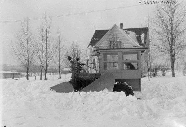 Side view of a truck with a snowplow on the front. The snowplow is plowing deep snow. There is a house in the background.