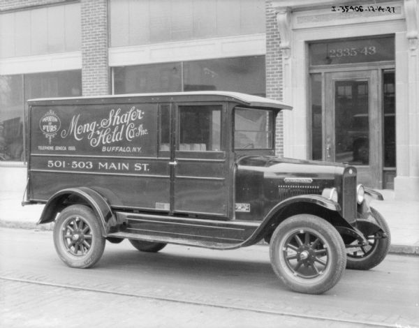 View across street towards a delivery truck parked along the opposite curb. There is a building with show windows in the background. The sign painted on the side of the truck reads: "Imported Furs. Meng-Shafer-Held Co. Inc."