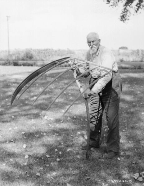 A man is posing outdoors holding an old reaping cradle.
