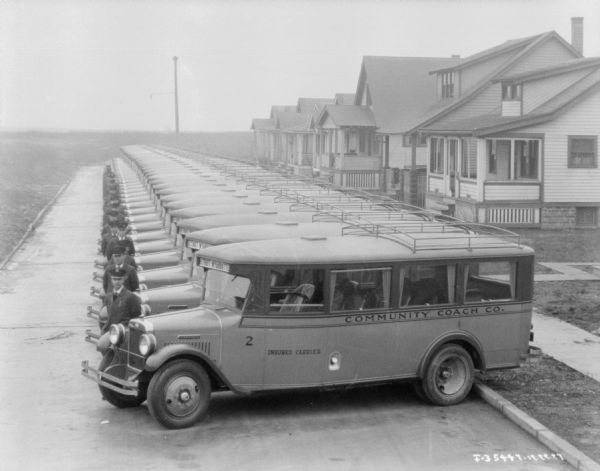 Elevated view of a fleet of buses (20-30) parked at a slight angle against a curb on the right, in a long row with an open field in the far background. The male chauffeurs are standing at the front right side of each bus. Houses are along the right side of the street.