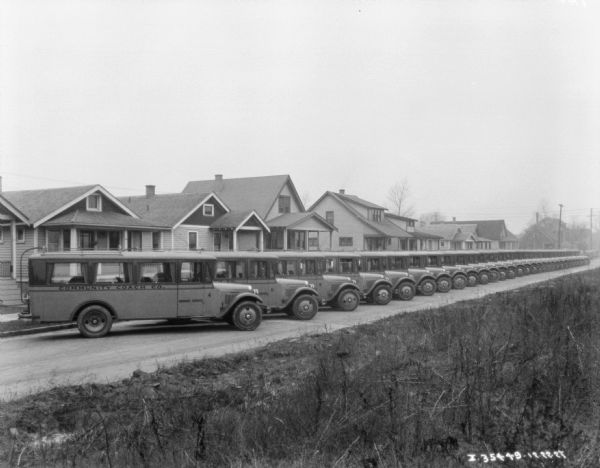 View from side of street towards a fleet of buses (20-30) parked at an angle against a curb in front of a row of houses.