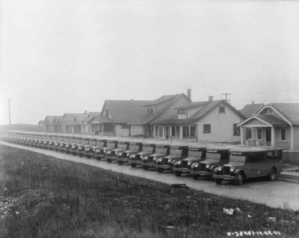 Slightly elevated view from field towards a fleet of buses (20-30) parked at an angle against a curb in front of a row of houses.