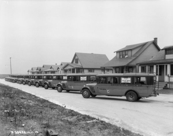 View from side of street towards a fleet of buses (20-30) parked at an angle against a curb in front of a row of houses. Chauffeurs are sitting in the passenger seat of each bus. Signs on the bus window read: "Body Built by Lang Body COmpany."