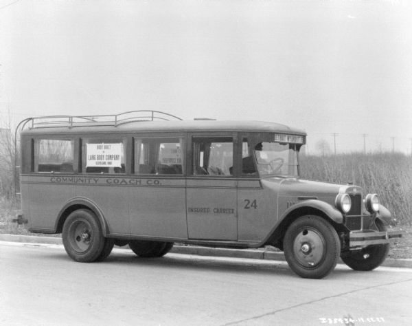 View across street towards a man sitting in the driver's seat of a bus.   The sign on the bus window reads: "Body Built by Lang Body Company."