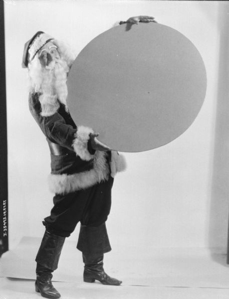 A man dressed in a Santa Claus costume is standing and holding a large disk in front of a white backdrop.