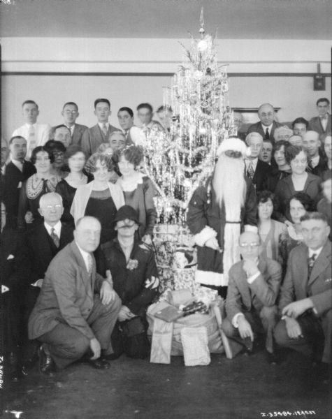 Group of people posing around a decorated Christmas tree, including a man in a Santa Claus costume.