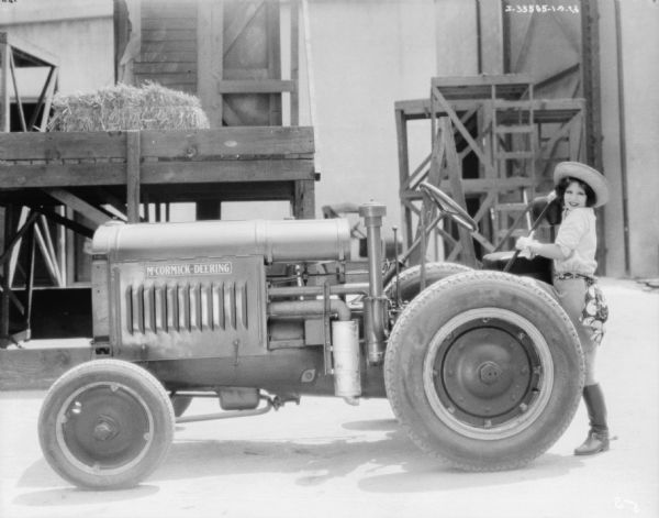 A woman is standing at the back of a McCormick-Deering tractor. She is smiling and is holding on to the tractor seat. The woman is wearing a straw hat, jodphurs and boots with spurs.
