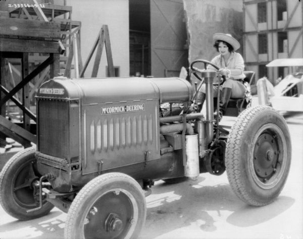 A woman is sitting in the tractor seat of a McCormick-Deering tractor. She is smiling and is holding on to the steering wheel. The woman is wearing a straw hat, jodphurs and boots with spurs.