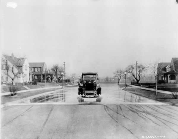A man sitting in the driver's seat of an International truck as a street cleaner is driving down the center of a paved street. The vehicle has a tank on the back and is spraying the street with water. There are houses on both sides of the street which is lined with trees and lampposts. Electrical towers are in background.