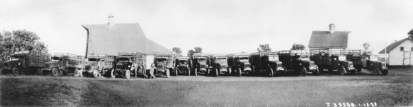 Panoramic view of a group of delivery trucks backed up to buildings. Men are standing near the trucks, which have high stake bodies in the truck beds for livestock.