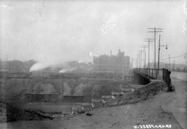 View from side of bridge, looking across towards a railroad train passing beneath the bridge along the opposite side of the bank. Behind the train is a large building that may be a dealership.