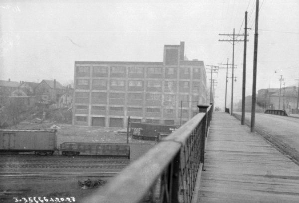 View down bridge railing towards a large dealership on the opposite bank. There are railroad cars on railroad tracks below the building.