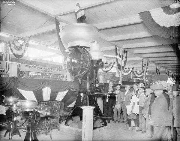 A group of men and women are looking at a giant cream separator on display in an exhibition hall. A newsboy is holding a stack of "Milwaukee Journal's."