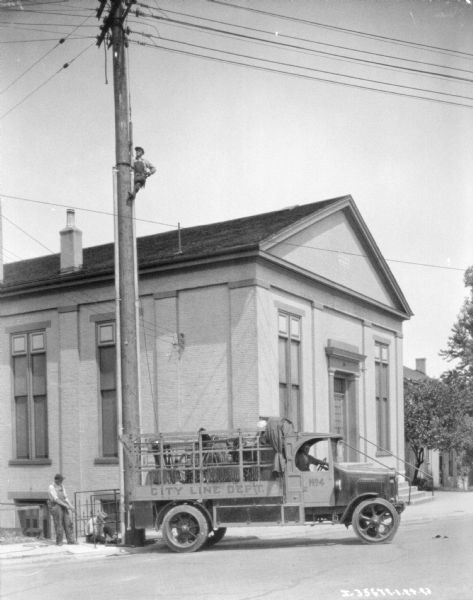 View across street towards men working around a City Line Dept. truck. One man is sitting in the driver's seat of the truck. Another man is halfway up a power line pole.