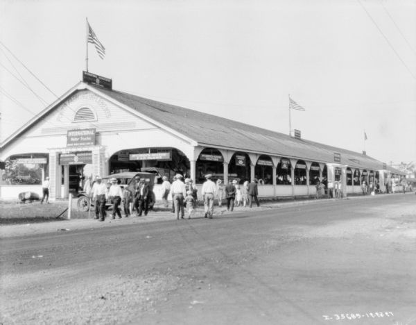 View across road towards a crowd walking towards a pavilion with McCormick-Deering International Harvester machines.