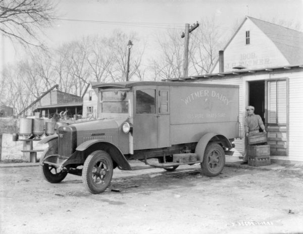 A man is loading crates into the back of a delivery truck backed up to a building. The sign painted on the side of the truck reads: "Witmer Dairy, Its Pure. That's Sure."