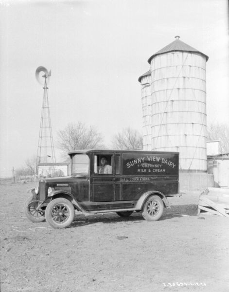 A man is sitting in the driver's seat of a milk delivery truck. A windmill, farm buildings and silo's are in the background. The sign painted on the truck reads: "Sunny View Dairy, Guernsey, Milk & Cream."