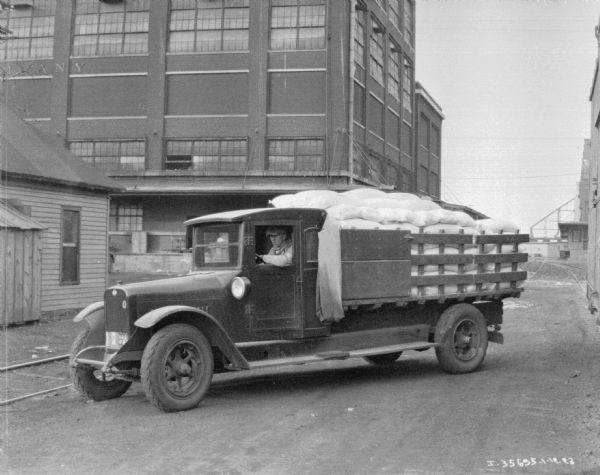 A man is sitting in the driver's seat of a delivery truck. The back of the truck has a stake body, and is piled high with filled sacks. In the background are industrial buildings.