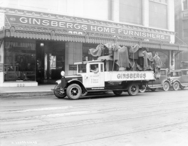 View across street towards a Ginsbergs Furniture delivery truck. There is a man sitting in the driver's seat, and another man standing at the back of the truck loaded with furniture. The Ginsbergs furniture store is in the background.