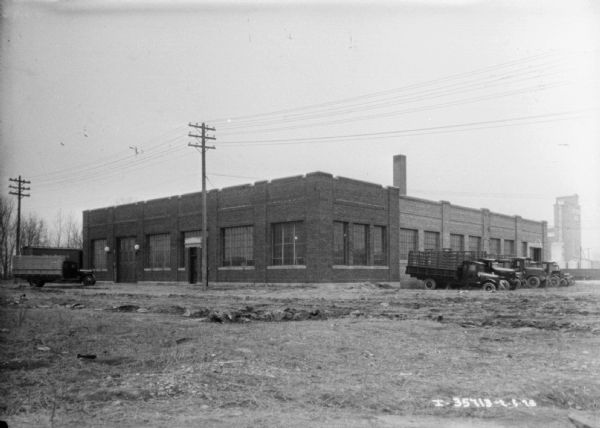 View across empty lot towards a dealership. Trucks are parked near the building. There is a large, industrial building in the far background.