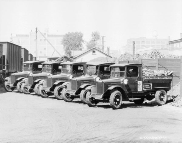 View across yard towards a fleet of six trucks parked at an angle. In the background is a large pile of coal. There are industrial buildings in the background. The signs painted on the trucks read: "Carbon Coal Co.," and "Carbon Chunks."