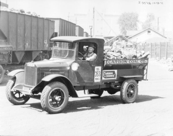 Three-quarter view from front left of a man sitting in the driver's seat of a coal delivery truck. The signs painted on the trucks read: "Carbon Coal Co.," and "Carbon Chunks."