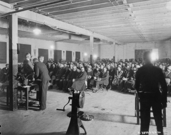 Men are standing near the front of a room facing an audience sitting in rows of chairs at a dealership.