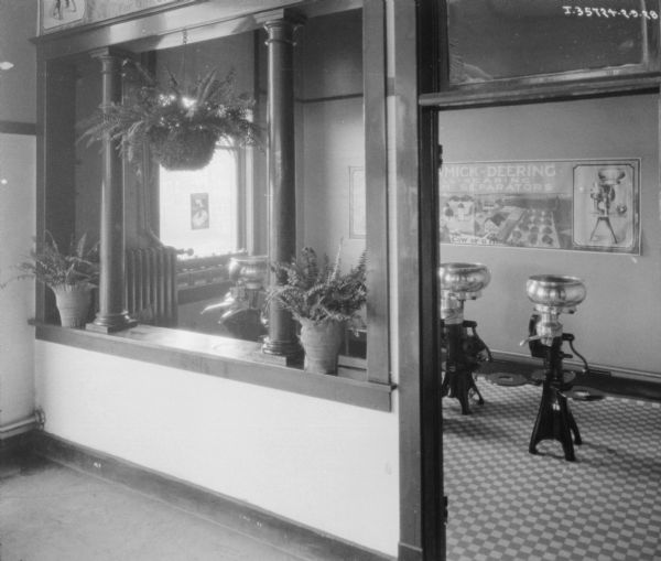 View looking towards a room in a dealership where cream separators are on display. There is a large McCormick-Deering Ball-Bearing Cream Separator poster on the wall in the background.