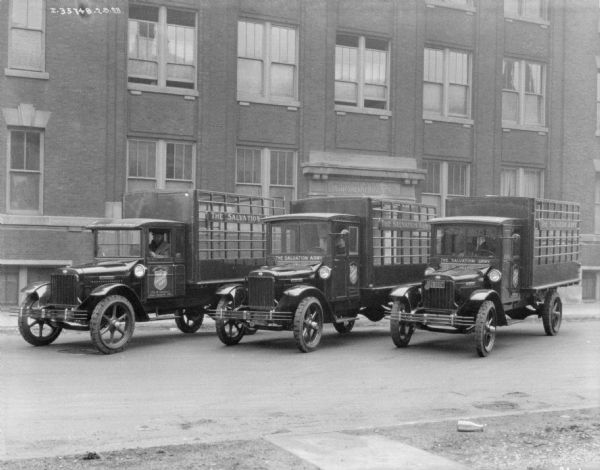 View of three trucks parked in a row at an angle. Men are sitting in the driver's seat of each truck. The Salvation Army building is in the background.