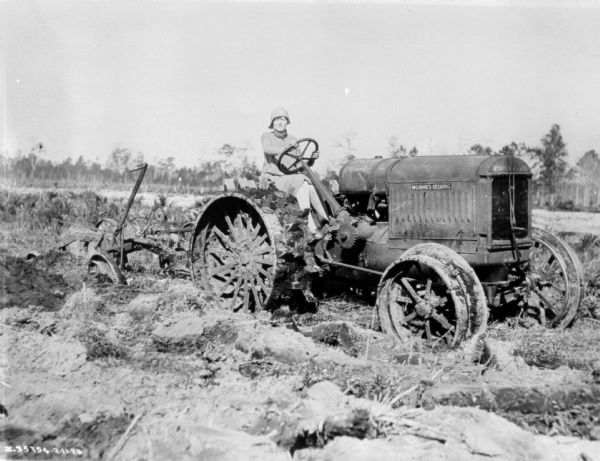 A woman wearing a dress and stylish hat is driving a tractor in a field, pulling what may be a plow.