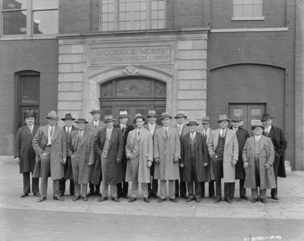 Group of male employees posing outdoors at McCormick Works. Behind the group is an entrance with a sign above the door that reads: "McCormick Works International Harvester Company."