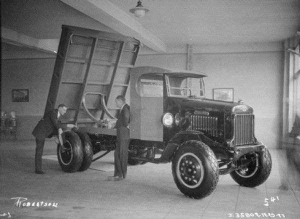A salesman is showing a client a truck inside a showroom. The truck bed is raised up.