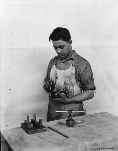 A man is repairing a part he is holding in his hand. He is wearing an apron and is standing behind a worktable. There is a white backdrop behind.