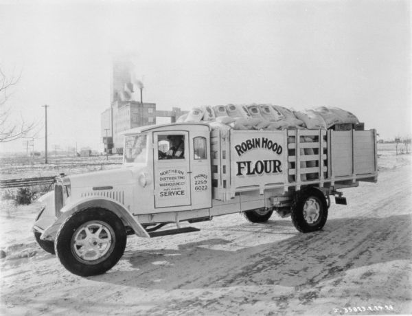 A man is sitting in the driver's seat of a delivery truck for Robin Hood Flour. The truck bed has a stake body, and is filled to the top with sacks of flour. A large industrial building is in the background.