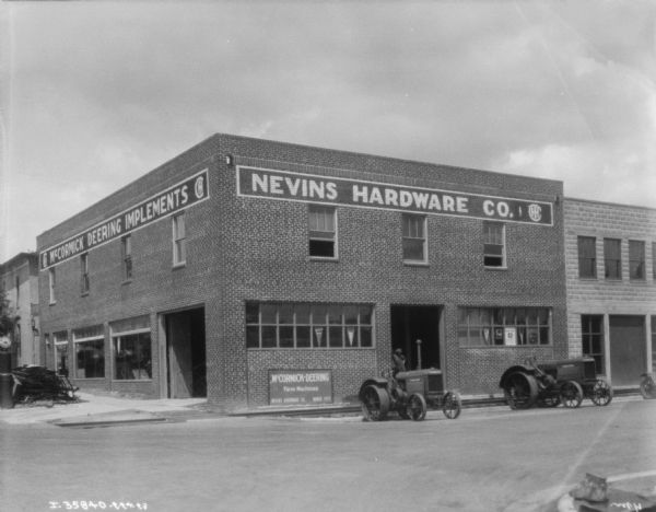 View across street towards a dealership on a corner. Tractors are parked along the curb. The sign on the building reads: "Nevins Hardware Co."