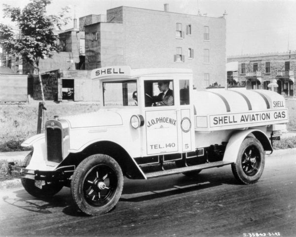 View across street towards a man sitting in the driver's seat of a Shell Oil delivery truck. In the background are commercial or apartment buildings. Sign on the side of the truck reads: "Shell Aviation Gas."