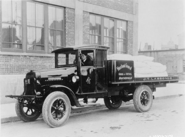 View across street towards a man sitting in the driver's seat of a truck parked along a curb in front of a brick building. The sign painted on the truck reads: "Marchand General, Gros et Detail, Boucherville," and "J.T. Lassonde."