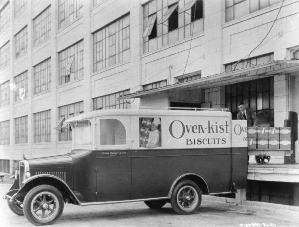 Delivery truck parked at a loading dock. The sign painted on the side of the truck reads: "Oven-kist Biscuits." A man is standing on the loading dock with a cart.