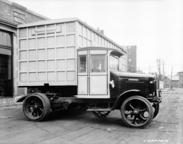 A man is sitting in the driver's seat of a bakery delivery truck parked outside of a brick building. The cab of the truck is at a right angle to the trailer, pointing towards the right, and the front of the trailer section of the truck is exposed. The sign on the front of the trailer reads: "Uneeda Bakers."