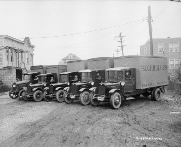 Five men are sitting in the driver's seats of five trucks parked in a row. The signs painted on the side of the trucks reads: "Buchmuller."