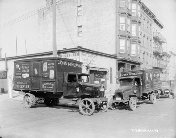 View across intersection of two Minervini Grocery trucks parked facing each other at the corner along a sidewalk in front of a storefront for Hoboken Auto Supply. The signs painted on the truck read: "John Minervini, Wholesale Groceries." A man is sitting in the driver's seat of the truck on the left, and a young boy is sitting in the driver's seat of the truck on the right.