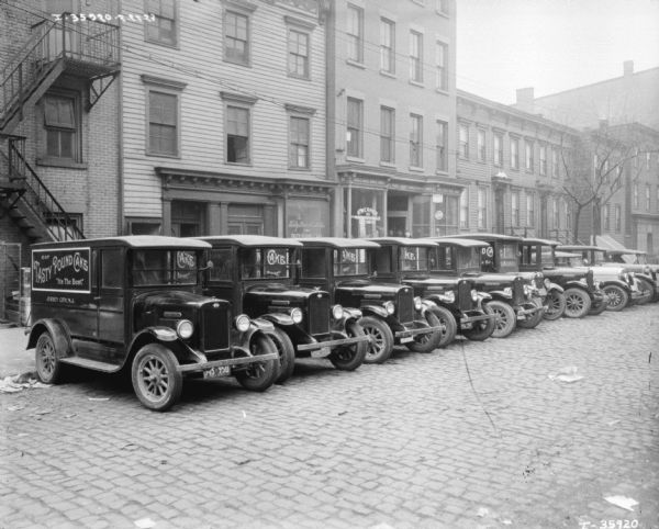 A fleet of trucks parked in a row in front of storefronts. The signs painted on the side of the trucks reads: "Eat Tasty Pound Cake, 'It's The Best.'"