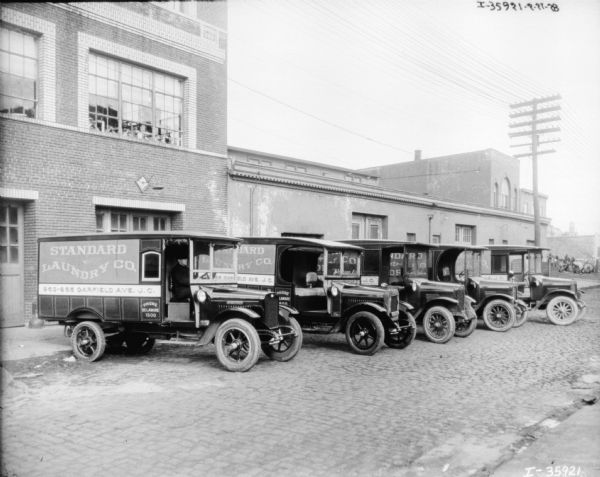 View down cobblestone street towards a fleet of five trucks parked in a row in front of a brick building.
