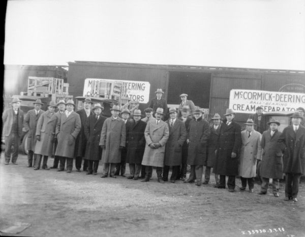 A group of men are standing in front of crates stacked in front of boxcars. The sign on the side of the railroad cars reads: "McCormick-Deering Ball-Bearing Cream Separators."