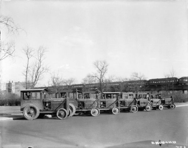 A group of men are posing in the driver's seats of McCormick-Deering tractors with enclosed cabs. The tractors are parked in a line at an angle against a curb. A train is on a railroad bridge in the background. The sign painted on the side of the tractors reads: "West Parks."
