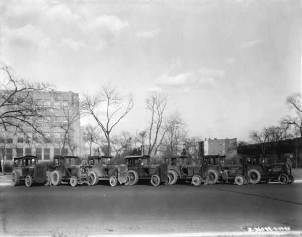 A group of men are posing in the driver's seats of McCormick-Deering tractors with enclosed cabs. The tractors are parked in a line at an angle against a curb. A train is on a railroad bridge in the background. The sign painted on the side of the tractors reads: "West Parks."